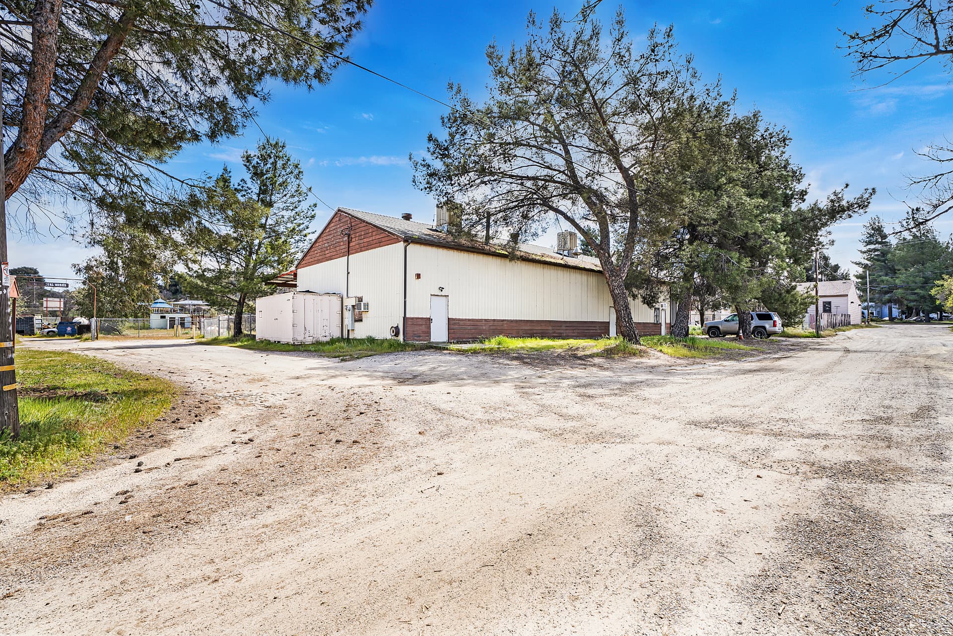 Large warehouse building shaded by pines at a dirt crossroads